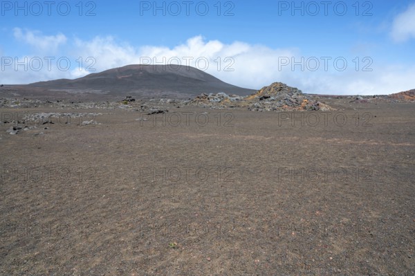 The Route du Volcan crosses the vast, rust-red volcanic desert of the Plaine des Sables, whose surface is dominated at extreme close angles by lava granules and sparse, barren grasses, while in the background a black-brown volcanic cone rises steeply into the blue sky under white cloud banks, Plaine des Sables, Highlands, La Reunion, France