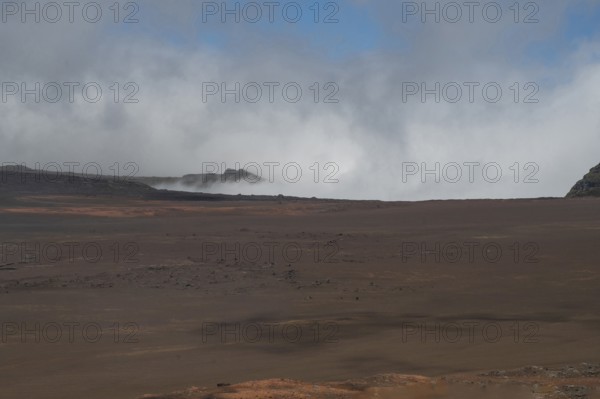 The Route du Volcan leads through the vast, rust-red lunar landscape of the Plaine des Sables, which is interspersed with dark lava rocks, while clouds hang over the eroded, steep volcanic rocks on the horizon, Plaine des Sables, Highlands, La Reunion, France