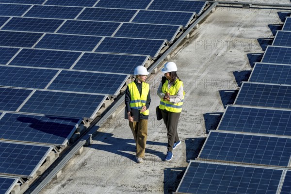Two women engineers wearing hard hats and high visibility vests walking and discussing work on a commercial rooftop with numerous solar panels. Symbolizing renewable energy and sustainable technology