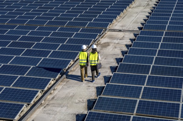 Two engineers, wearing hard hats and safety vests, walking between rows of solar panels on a rooftop, symbolizing renewable energy and sustainable power generation