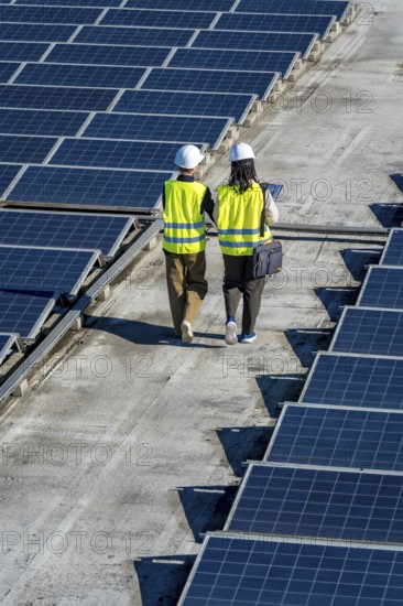 Two renewable energy engineers, wearing hard hats and reflective vests, walking along a rooftop solar power installation, performing maintenance and ensuring sustainable clean energy production