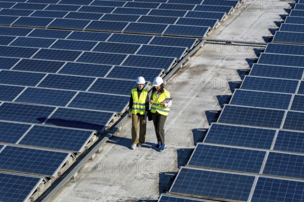 Two engineers, a man and a woman, are working outdoors, reviewing plans and inspecting rows of solar panels on a large rooftop array, embodying renewable energy and sustainability