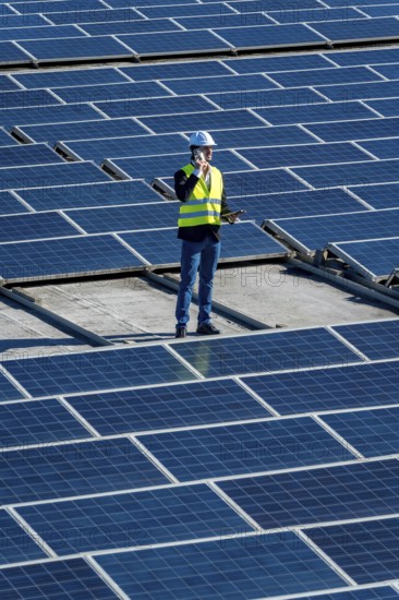 Engineer in hard hat and safety vest on commercial rooftop amid solar panels, using mobile phone while inspecting and maintaining photovoltaic array for clean, sustainable energy projects