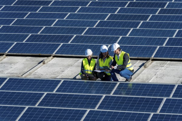 Team of engineers in hard hats and vests is discussing data on a tablet, working on a large solar panel installation on a rooftop during a sunny day