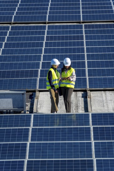 Female engineers wearing hard hats and high visibility vests discussing data on a digital tablet while standing on a rooftop solar panel installation, representing teamwork and renewable energy