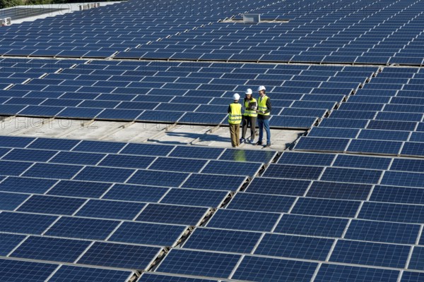 Engineers wearing safety vests and hard hats are standing on a large rooftop solar farm, reviewing plans and collaborating on a renewable energy project