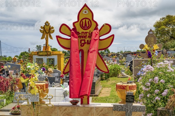 A bright red and yellow Hindu tombstone with religious symbols and a draped cloth stands as a sign of multi-religious culture and religious tolerance in the Saint-Gilles-les-Hauts cemetery in the midst of other graves, Saint-Gilles-les-Hauts, La Reunion, France