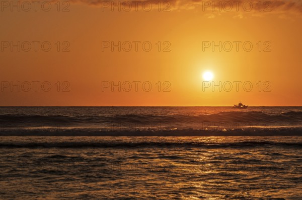 A warm sunset light bathes the sky over the Indian Ocean in orange and gold while a fishing trawler fishes on the dark waves in the ocean, Saint-Gilles, La Reunion, France