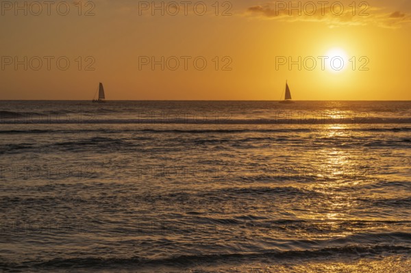 A warm sunset light bathes the sky over the Indian Ocean in orange and gold as two small sailboats cruise on the dark waves in the sea, Saint-Gilles, La Reunion, France