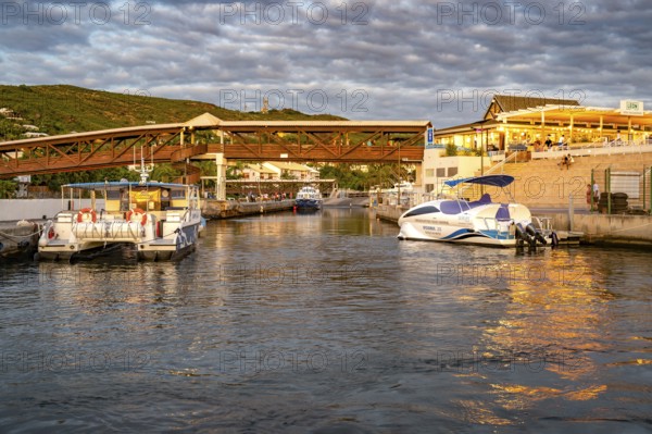 Several boats and catamarans are moored on the jetty in the port of Saint-Gilles, whose water reflects the warm reflections of sunset light and the surrounding restaurants and a wooden footbridge, Saint-Gilles, west coast of Reunin, Ile de la Reunion, France