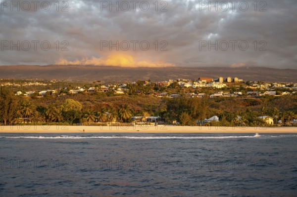 The bright sandy beach of the Saint-Gilles lagoon with bathers merges into a coastal landscape with palm trees and white houses, while the mountains are illuminated by warm evening light in the background and a jet ski ride through the sea, Saint-Gilles, La Reunion, France