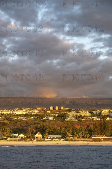 The bright sandy beach of the Saint-Gilles lagoon with bathers merges into a coastal landscape with palm trees and white houses, while the mountains are illuminated by warm evening light in the background, Saint-Gilles, La Reunion, France