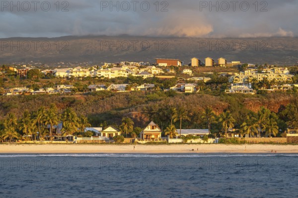 The bright sandy beach of the Saint-Gilles lagoon with bathers merges into a coastal landscape with palm trees and white houses, while the mountains are illuminated by warm evening light in the background, Saint-Gilles, La Reunion, France