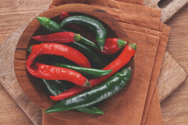 Vibrant red and green chili peppers rest in a wooden bowl, placed on a textured brown cloth atop a wooden table, creating a fresh and inviting scene