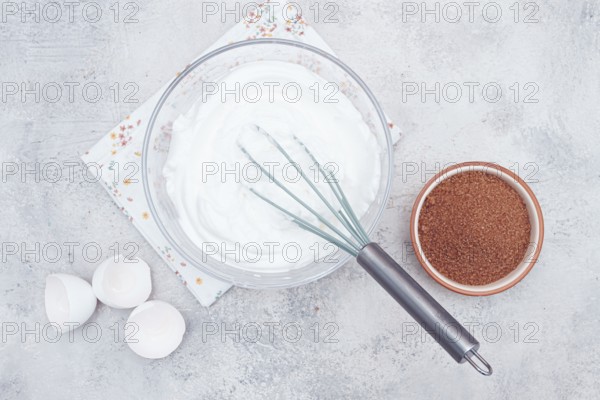 A clear bowl holds whipped egg whites, ready for dessert preparation. Next to it, a bowl of brown sugar and eggs sit on a floral napkin in a bright kitchen atmosphere