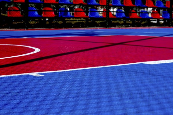 The basketball court features a striking red and blue design illuminated by bright sunlight. The court lines are clearly visible, creating a dynamic playing surface for sports activities
