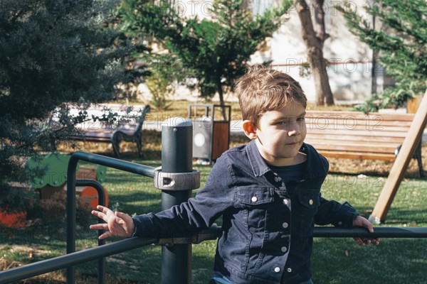 A young boy is sitting on a playground structure in a park, surrounded by trees and grass. He looks thoughtful and relaxed in the warm sunlight of the afternoon