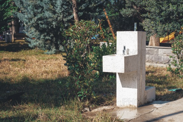 In a peaceful park, a modern water fountain is positioned among lush green plants and grass. The bright sunlight highlights its clean design, inviting visitors to enjoy fresh water