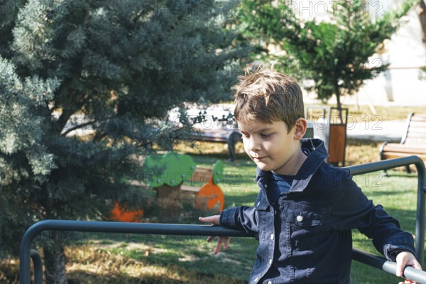 A young boy sits on a playground railing in a sunlit park, surrounded by greenery and trees. He looks thoughtful while enjoying his time outdoors