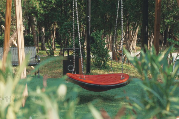 A vibrant red swing hangs suspended in a green park, surrounded by tall trees and wooden benches. The area appears peaceful with sunlight filtering through the leaves