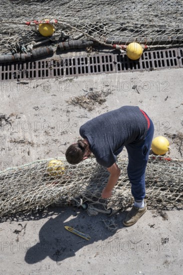 Seen from above, a fisherwoman in blue pants bends over a large fishing net spread out on the dry concrete for repair, the edges of which are marked with yellow floats and reflect the busy atmosphere of the harbor, Erquy, Saint-Brieuc Bay, Côtes-d'Armor, Brittany region, France
