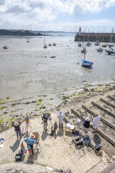 A group of plein-air amateur painters and artists stand with easels on a concrete-like ramp and looks at the large, muddy sandy beach, on which numerous sailing and fishing boats dry at low tide and in the background the pier with the small, red and white lighthouse marks the harbor entrance, Erquy, Saint-Brieuc Bay, Côtes-d'Armor region, France
