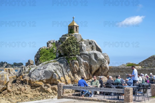 The small stone chapel of Saint-Gildas is perched on a distinctive rock of pink granite, while in the foreground a group of people relaxing on a café terrace in front of a stone wall and enjoying the bright sun, Port Blanc, Penvenan, Cotes-d'Armor department, Brittany region, France