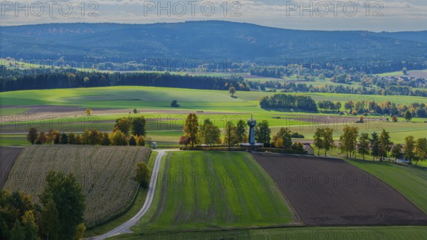 In the midst of a vast autumn landscape with green fields and colorful trees, there is a small observation tower. In the background, you can see rolling hills and a clear blue sky, the light is soft. Grenzland Tower, Bad Neualbenreuth, Upper Palatinate