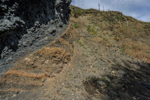 The scene shows different layers of earth and rock formations in a rural area on a clear day. Dry grass and small plants are visible. Železná hurka, Czech Republic