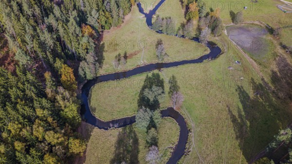 A clear river snakes through a green meadow, surrounded by colorful autumn trees. The picture shows the tranquil beauty of nature during autumn. Falkenberg, Upper Palatinate