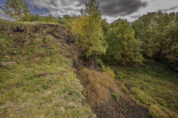 The gentle slope of an extinct volcano shows a variety of trees with colorful autumn leaves. The surrounding area is quiet and offers a beautiful view of green grass and wooded areas. Železná hurka, Czech Republic