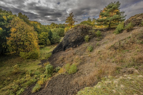 The scene shows a varied autumn landscape with colorful trees. The sky is cloudy and makes the colors of nature appear vivid. The slope of the extinct volcano is clearly formed. Železná hurka, Czech Republic