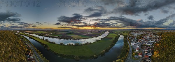The landscape shows a picturesque sunset over a river. A city is visible in the foreground, surrounded by green fields and clouds. Bogen, Lower Bavaria