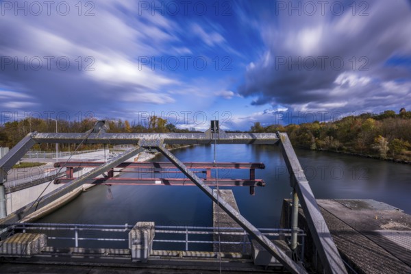 The view shows a lock on a calm body of water. The autumn colors of the trees are reflected in the water as clouds pass by quickly. Landau an der Isar, Lower Bavaria