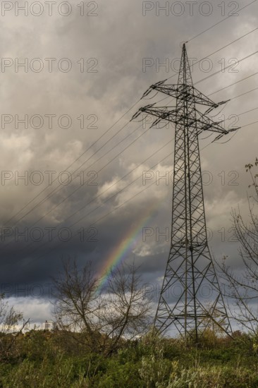 There is a high-voltage pylon in a rural area. Behind the mast, a rainbow appears in the sky. It is cloudy and looks like a thunderstorm. Bogen, Lower Bavaria