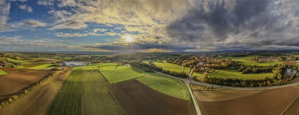 A vast landscape of green fields and brown fields under a dynamic sky. The sun sets behind the hills as the clouds thicken. Bogen, Lower Bavaria