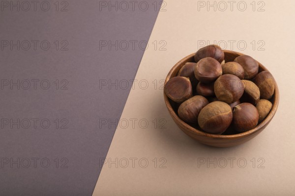 Wooden bowl with raw edible ?hestnuts on brown and gray paper pastel background, side view, copy space, minimalism