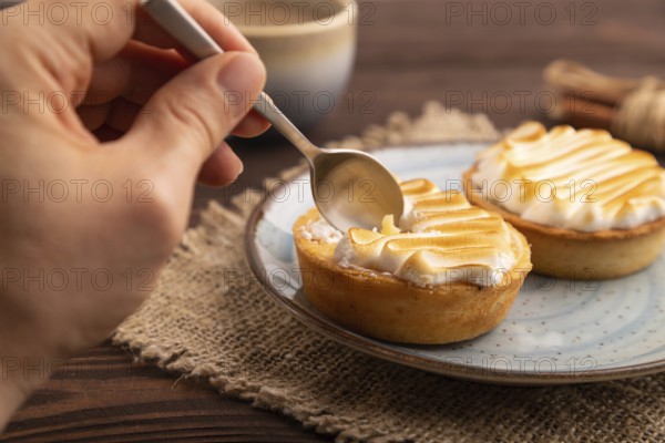 French lemon tart with meringue with hand on brown wooden background, cup of coffee, linen textile, side view, close up, selective focus