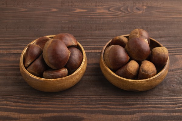 Wooden bowl with raw edible ?hestnuts on brown wooden background, side view, close up, minimalism