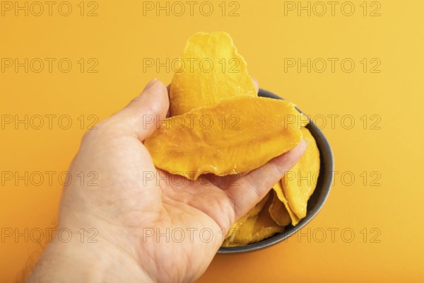 Dried Mango in blue ceramic bowl with hand on orange pastel paper background. Side view, close up. healthy food, minimalism