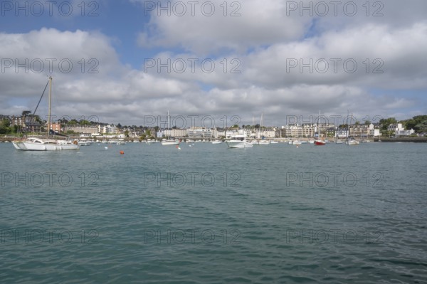 A series of small sailboats, probably optimists, with red and white ringed sails, move in formation parallel to the coast across the slightly curled turquoise water, behind which a beacon can be seen on a rocky headland, Plerin, Saint-Brieuc Bay, Cotes-d-Armor, Brittany, France