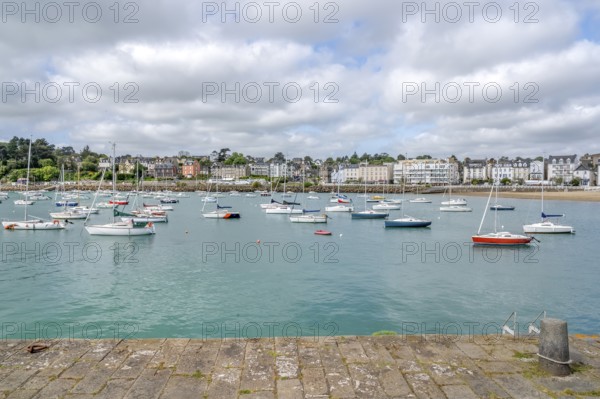 The view from a stone harbor wall shows numerous sailing and motor boats on the turquoise water of the harbor basin, behind which the dense, multi-storey development of the seaside resort stretches along the coastline, Saint-Quay-Portrieux, Cotes-d'Armor region, France