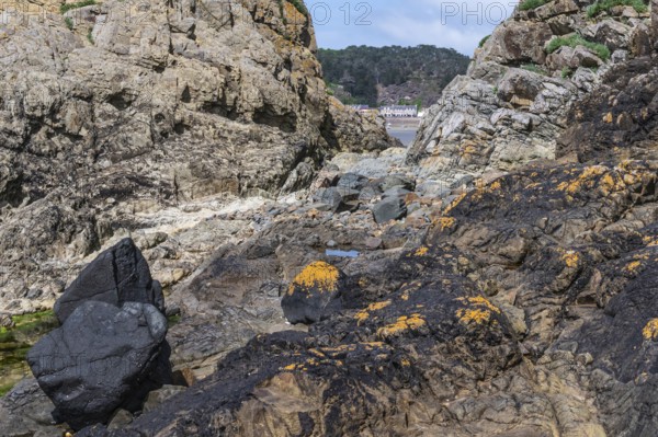 A rugged rock formation, dark and yellow-orange covered with lichens, which forms a narrow opening through which the wide sandy beach and the buildings of the town of Erquy can be seen in the distance, Erquy, Saint-Brieuc Bay, Côtes-d'Armor, Brittany region, France