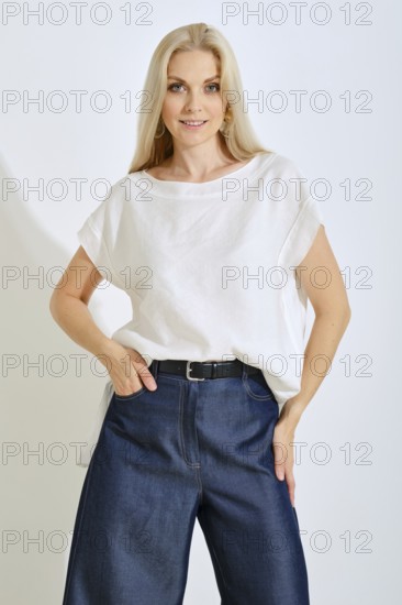 A blonde woman stands confidently in a bright studio, wearing a simple white top and loose blue denim pants