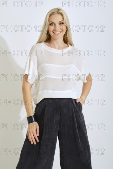 A model is wearing a stylish white top with horizontal stripes and dark, wide-legged pants. She smiles confidently, showcasing modern fashion in a bright, minimal setting