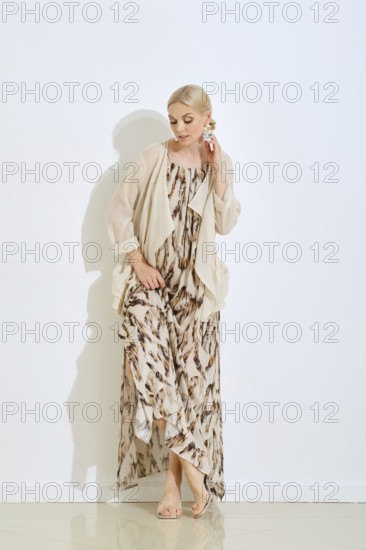 A woman showcases a fashionable long dress with a unique pattern. She stands elegantly by a wall, wearing a light shawl and stylish footwear, highlighting her refined style and poise