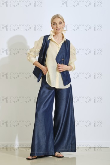 A model showcases a fashionable outfit with a denim vest and wide-leg pants paired with a light blouse. The background is bright and simple, enhancing the modern look of the ensemble
