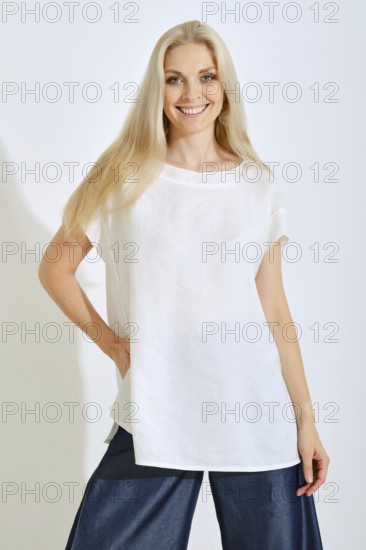 A woman stands smiling with long, light hair, dressed in a simple white top and dark denim pants. She looks relaxed and confident against a plain white background