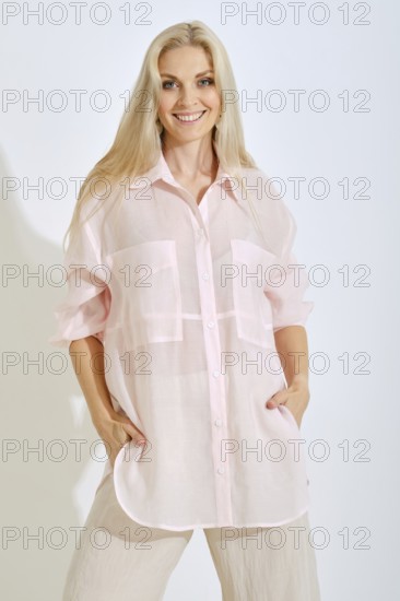 A woman stands confidently in a bright studio, wearing a light pink oversized shirt with pockets and beige pants