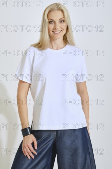 A woman stands confidently, wearing a plain white t-shirt paired with loose denim pants. She smiles warmly, with soft lighting highlighting her features in a bright indoor environment